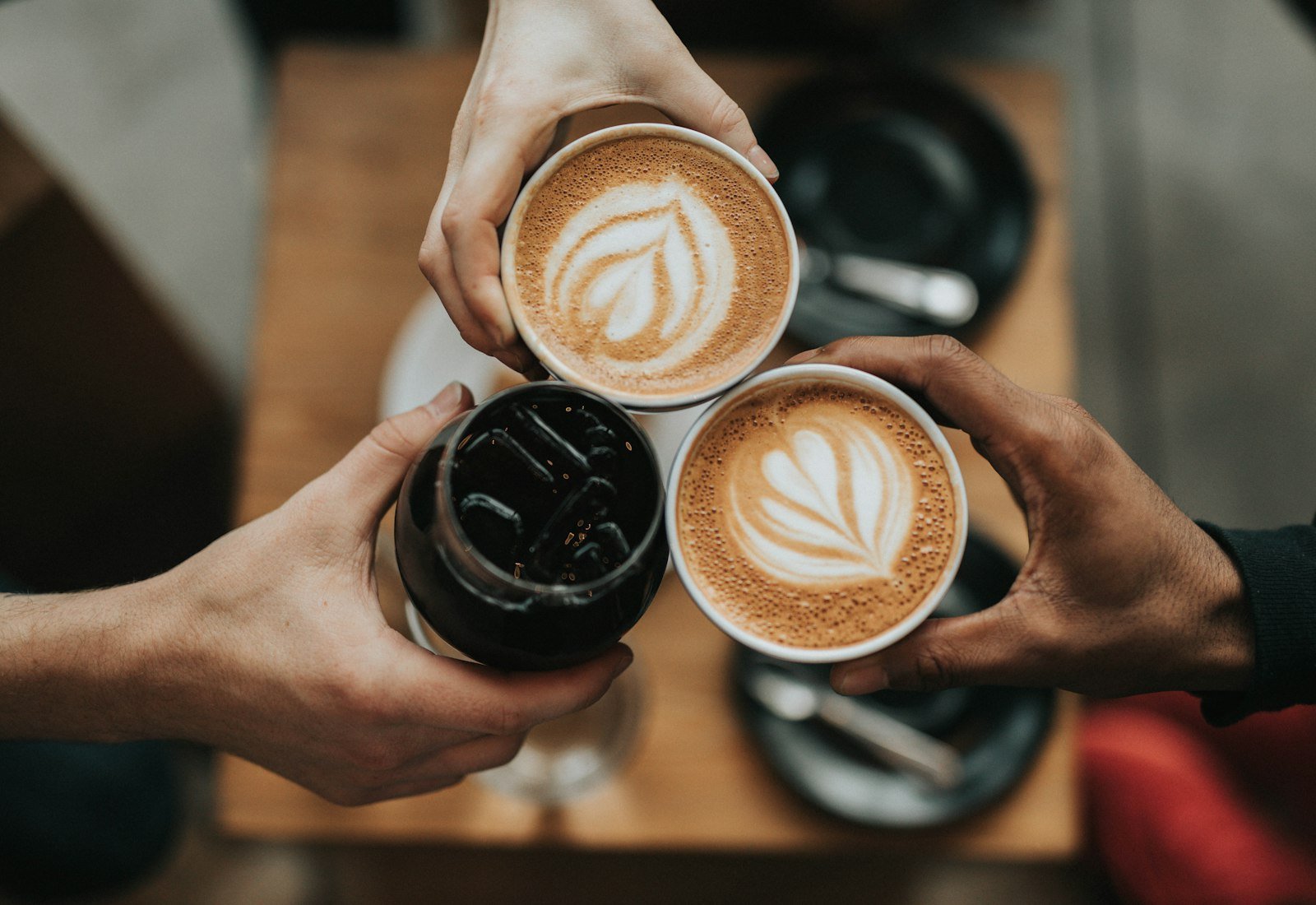 Three hands raising two lattes and a dark drink together over a cafe table